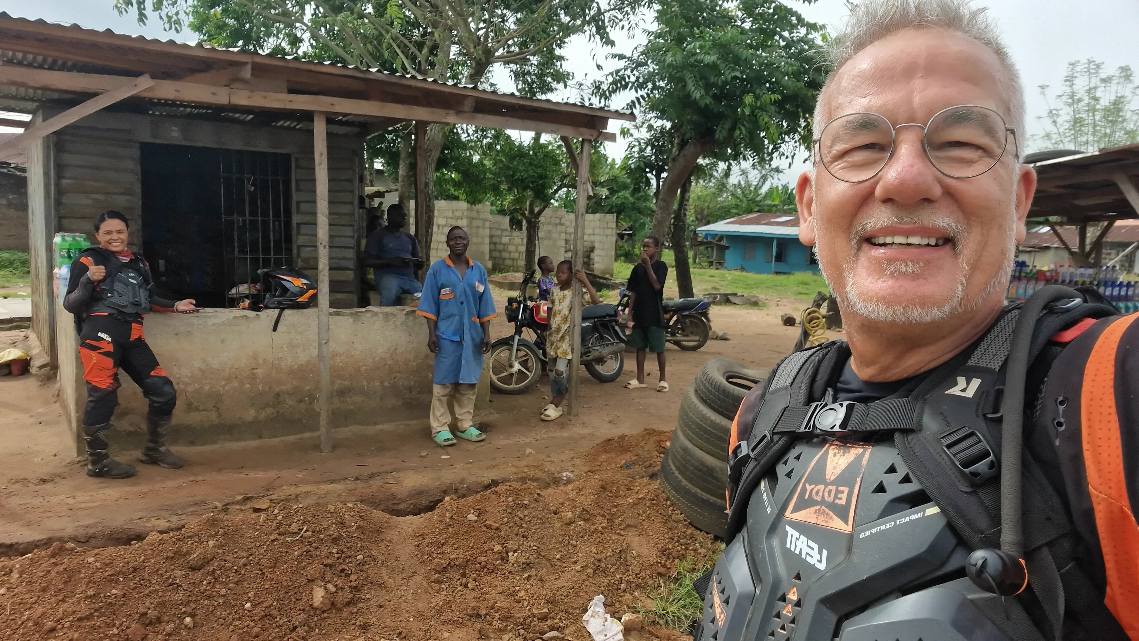 Eddy takes a selfie in body armour at a roadside workshop, Max laughing in riding gear behind him beside a parked motorcycle and a group of onlookers.