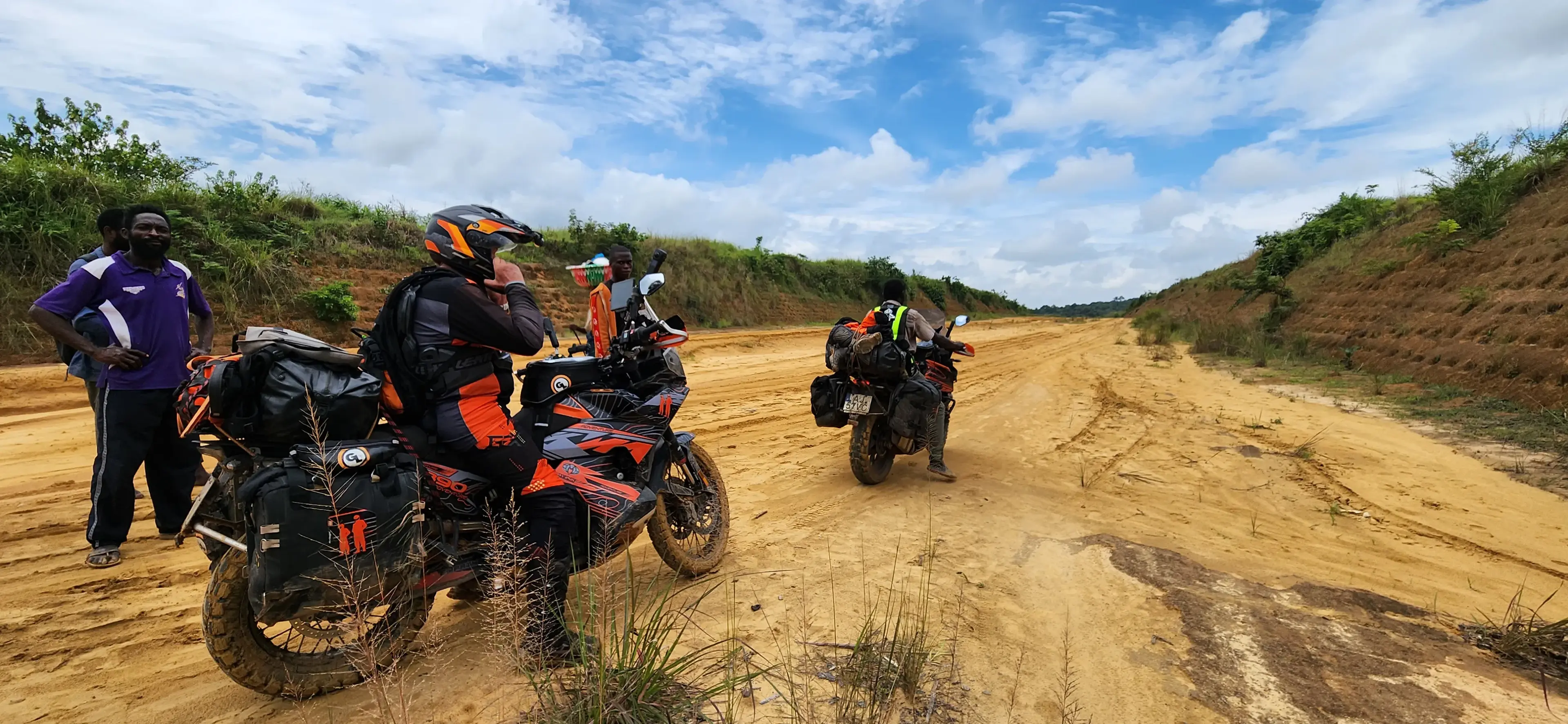 Two loaded adventure motorcycles resting on a sandy track under a wide, cloud-scattered sky.