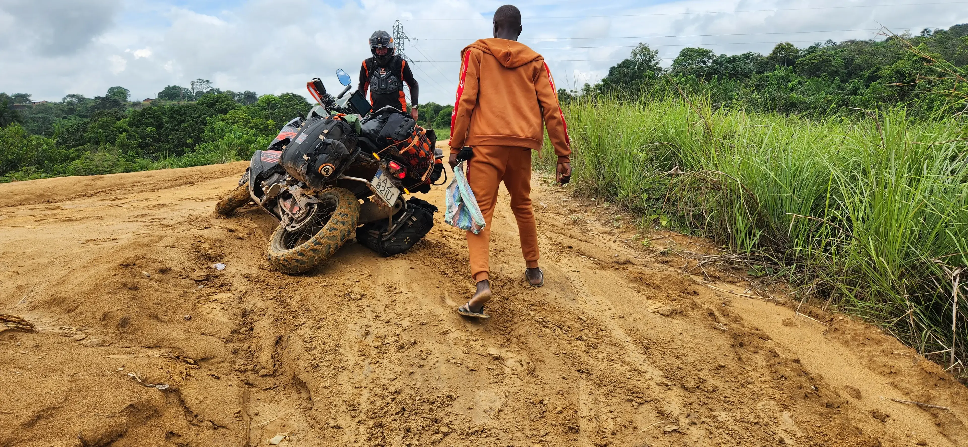 A loaded adventure motorcycle stuck in deep red mud on a dirt road, the rider in gear standing beside it while a man in an orange tracksuit walks over to help, tropical scrub on either side.
