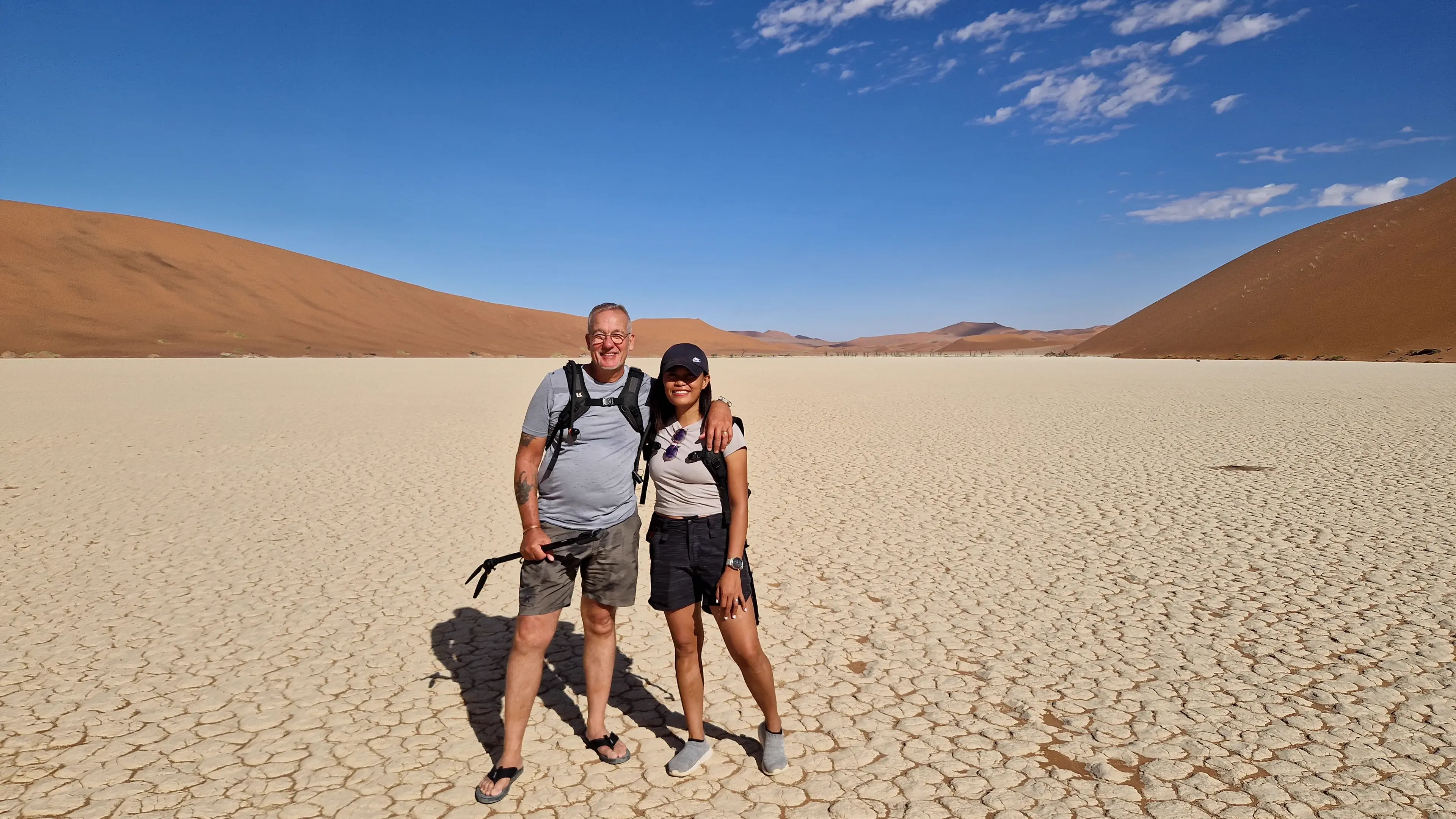 Eddy and Max standing together on the pale cracked clay of a desert pan, red dunes rising behind them under a hard blue sky.