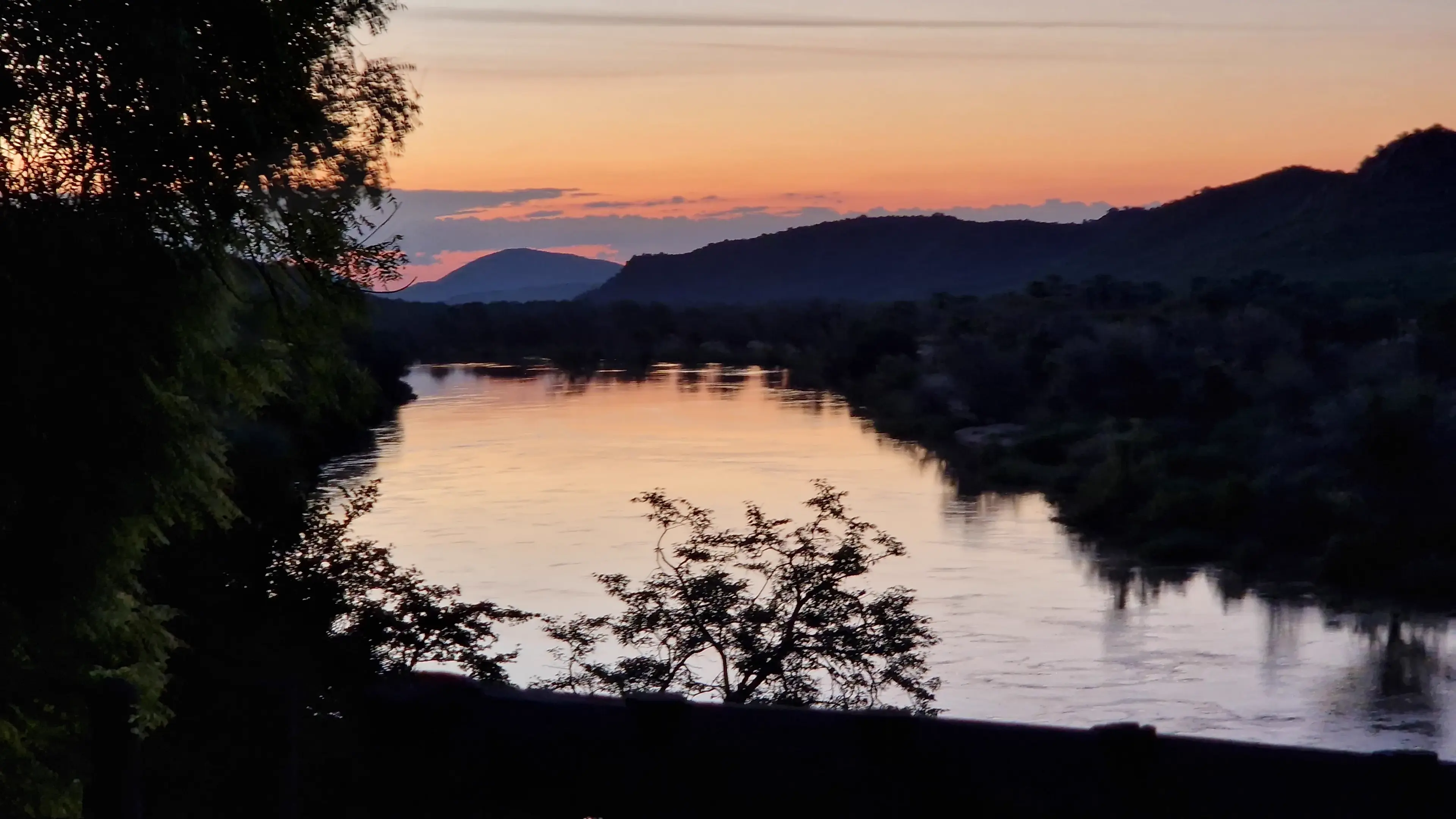 A wide river at dusk, low hills and silhouetted trees along the far bank under an orange sky.