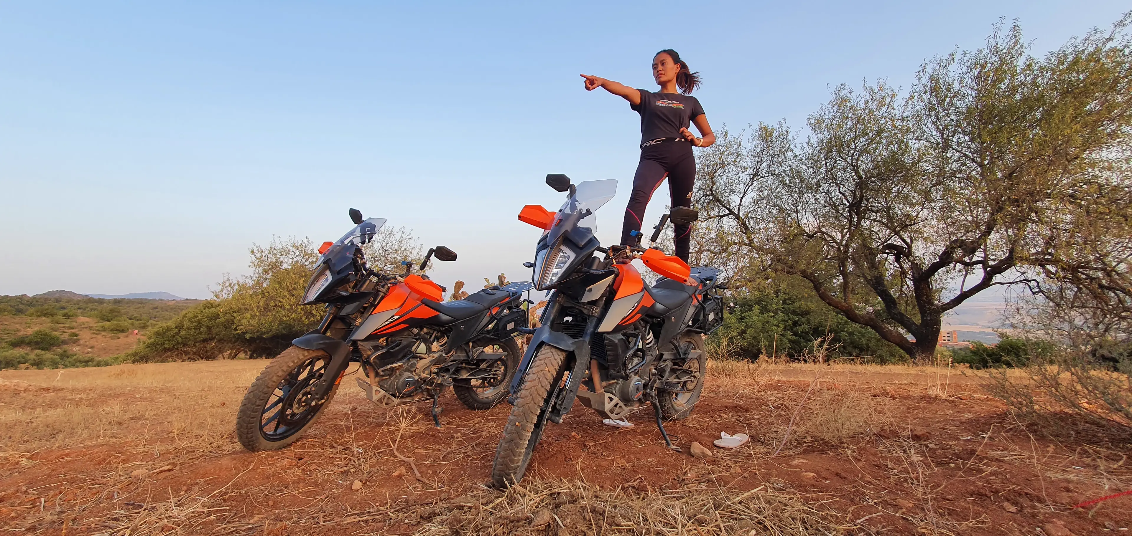 Two dusty orange adventure motorcycles parked on a dirt track at golden hour, a rider standing on one of them and pointing toward the horizon under a clear sky.