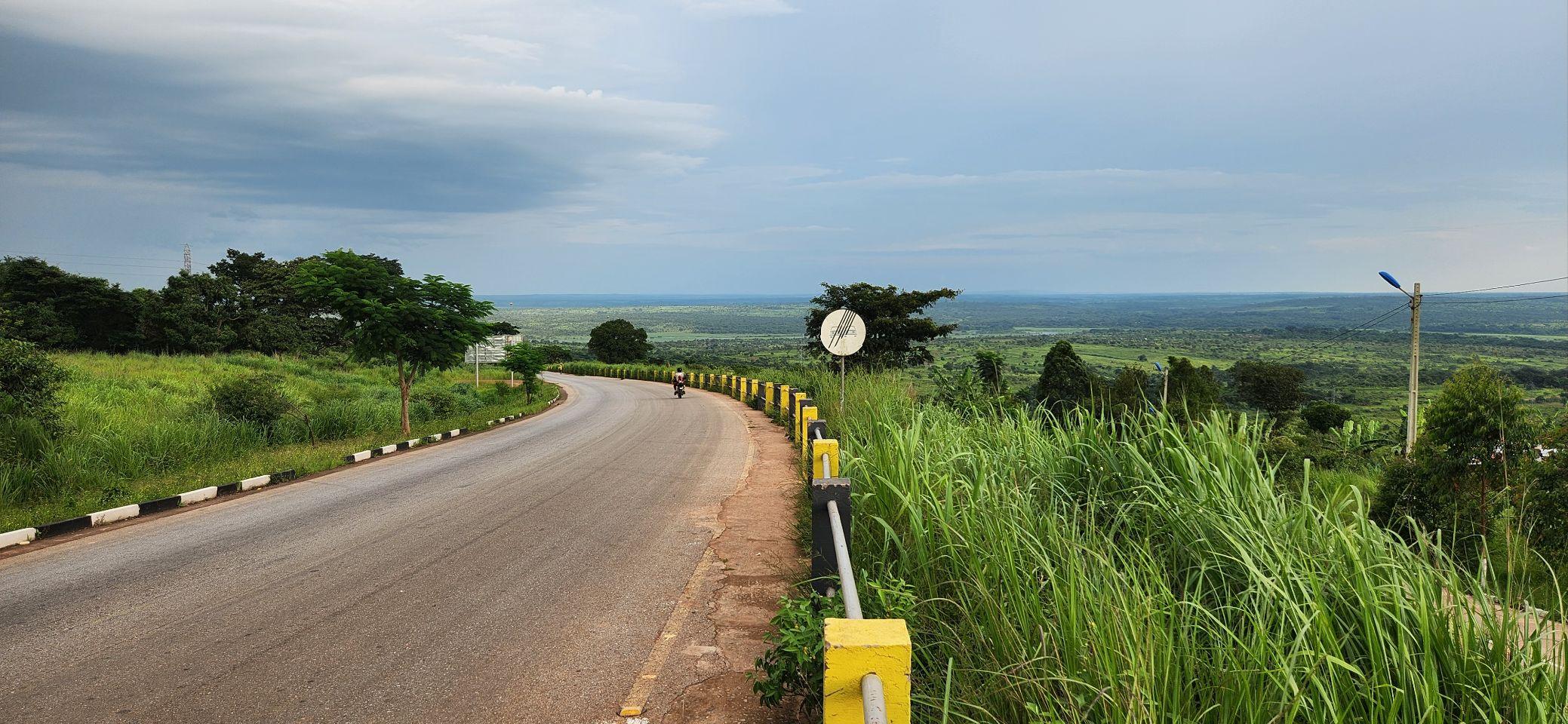 A quiet paved road curves through green hills toward Calandula, a lone motorcyclist visible in the distance.
