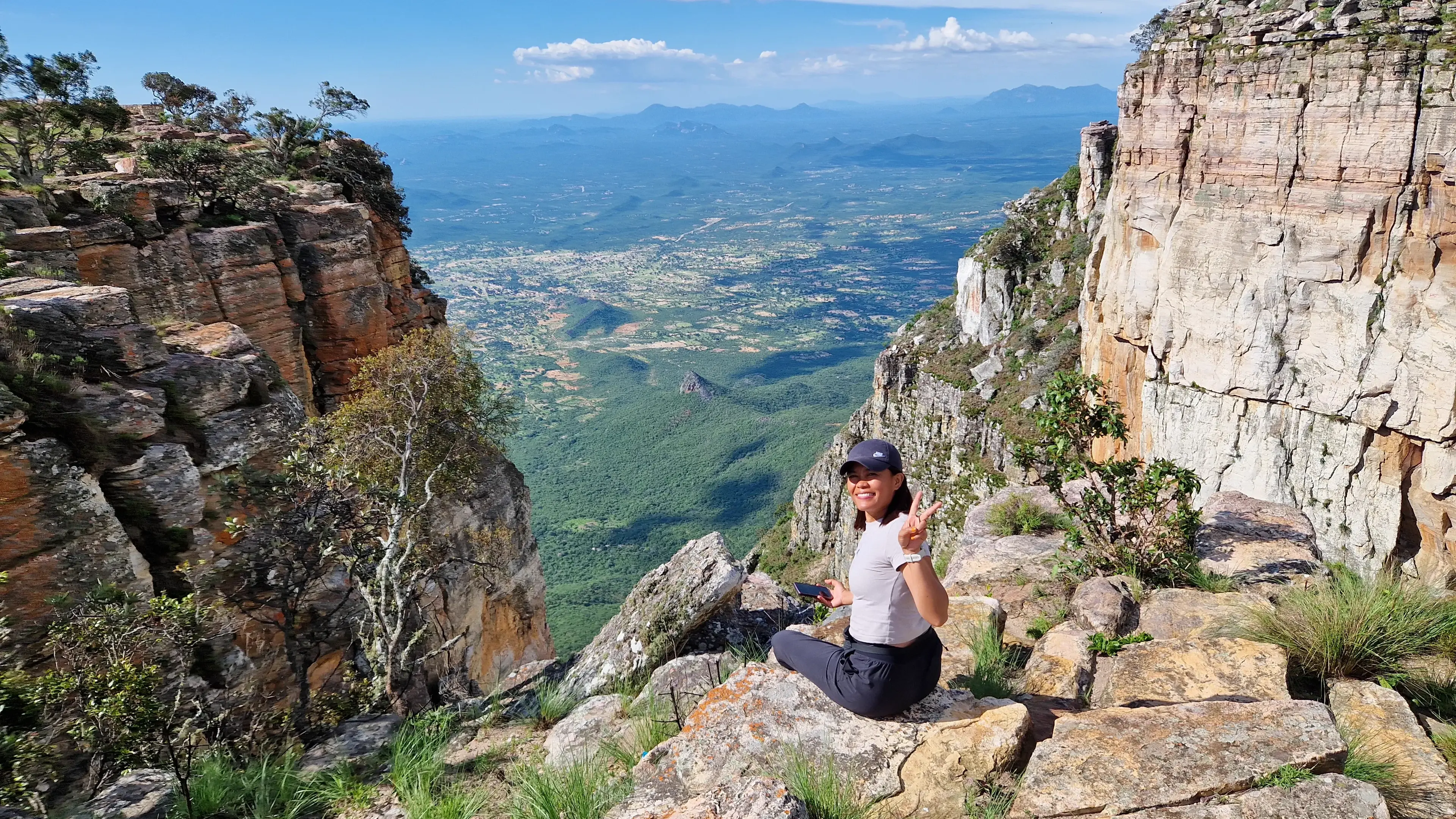 A person sitting on a rocky cliff edge, looking out over a wide green valley far below.