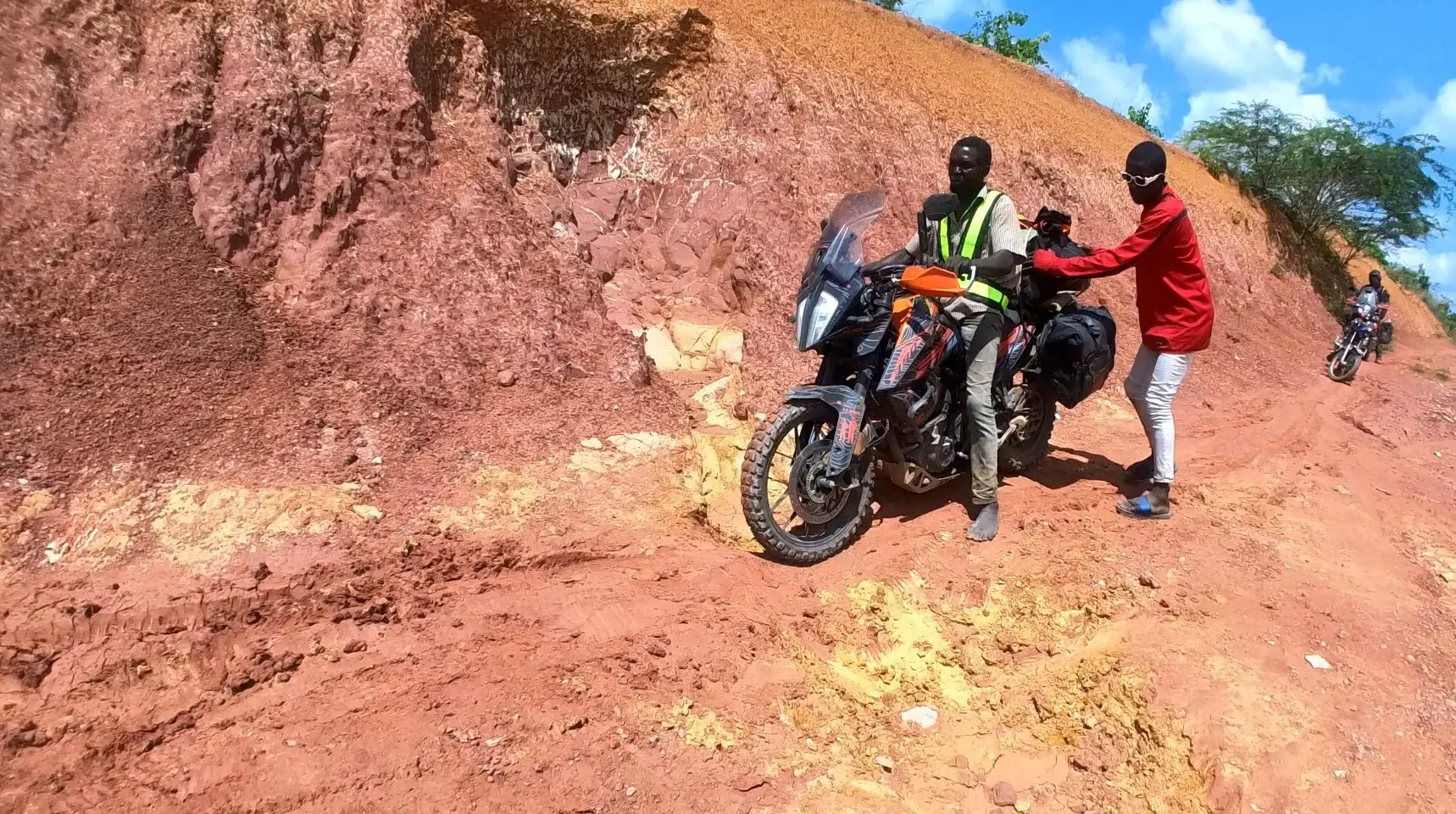 A local rider in an orange helmet helping push a loaded motorcycle through deep red mud.