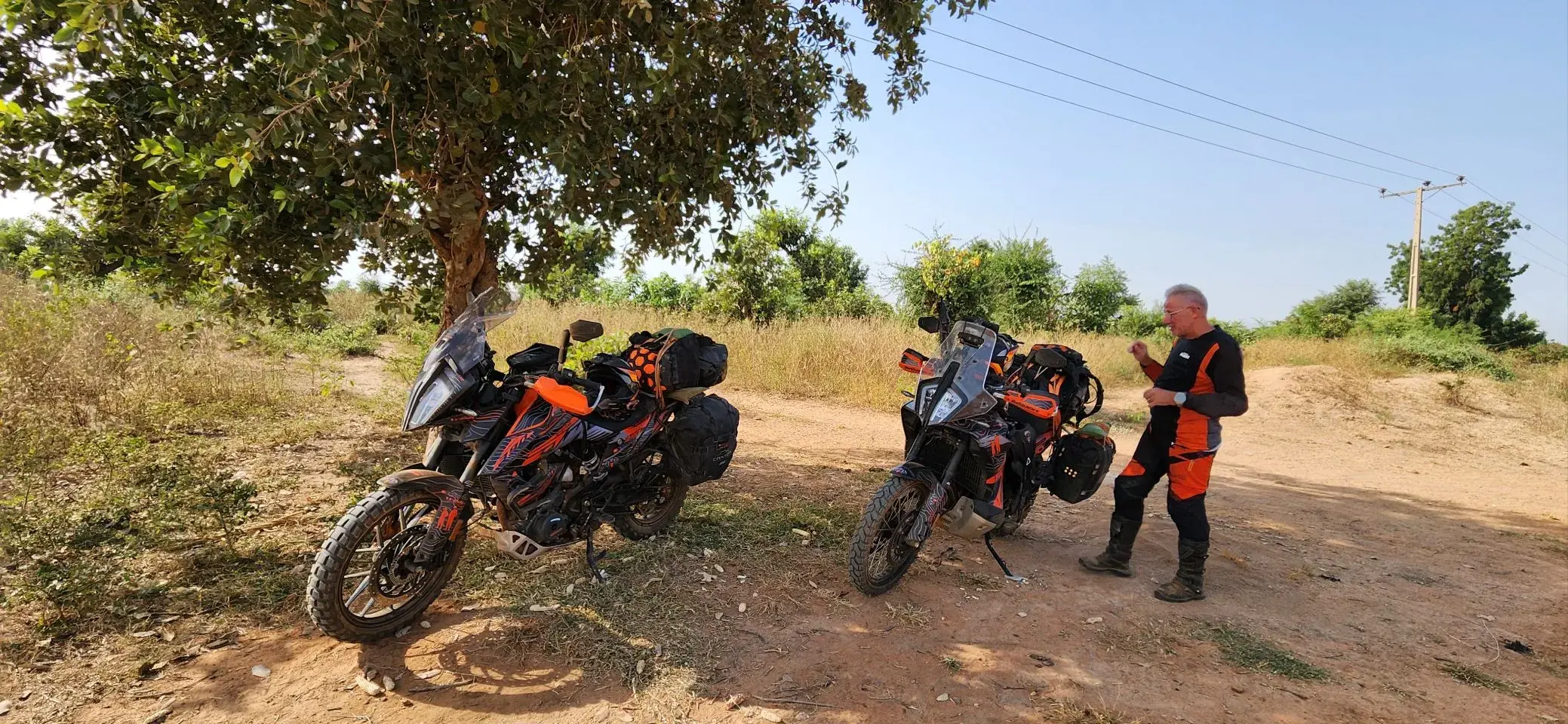 Two loaded orange and black KTM adventure motorcycles parked in the dappled shade of a large tree on a sandy red track, dry grass and scrub behind, with Eddy standing to one side in riding gear adjusting his jacket.