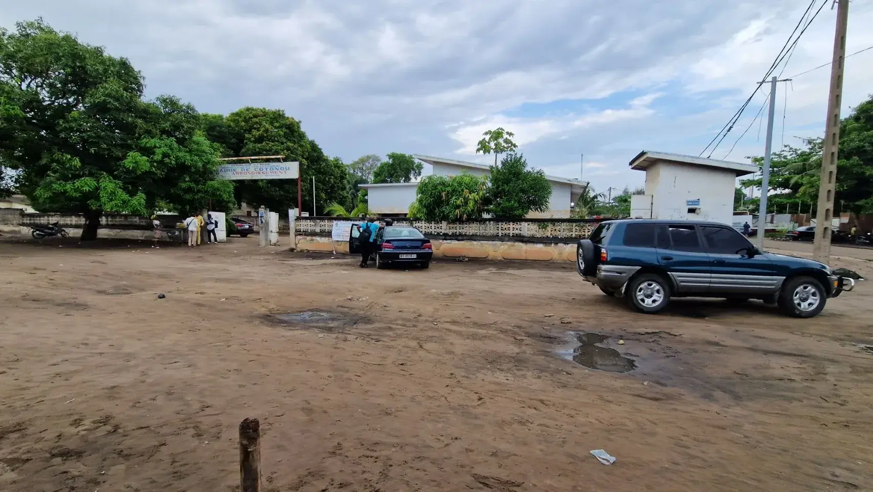 A rusted and cracked painted metal sign above a dirt driveway reading Republique du Benin, Departement du Littoral, Mairie de Cotonou, with a leafy tree overhanging and an administrative building with peeling paint behind it.