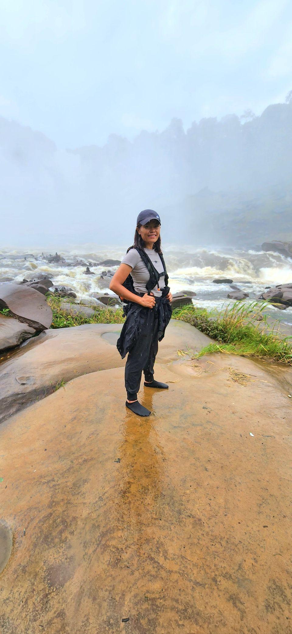 Max stands at the base of Calandula Falls in wet clothing, the churning river and misted rock face behind her.