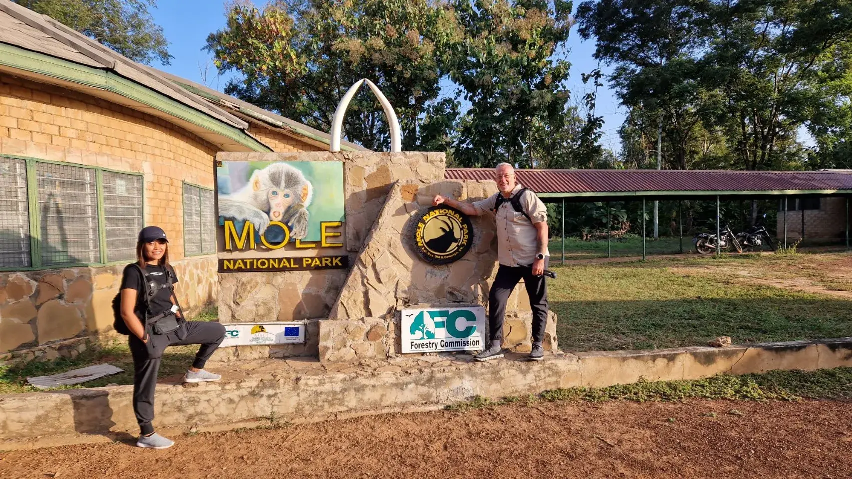 Max and Eddy stand either side of the stone Mole National Park entrance sign, which carries a painted baboon and a pair of white tusks, in low early morning light.
