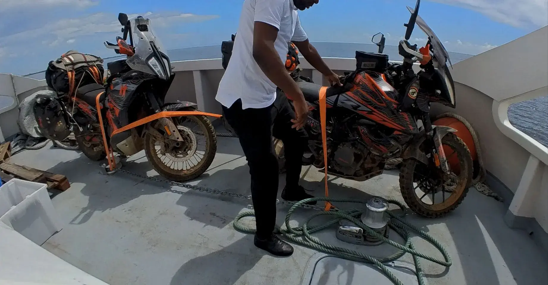Two adventure motorcycles strapped upright on the open deck of a boat at sea, a crew member leaning over to check a ratchet strap.