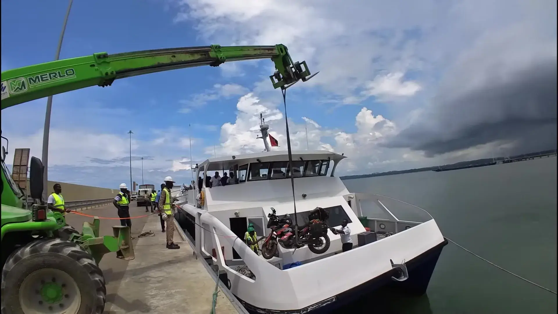 A green Merlo telehandler swings a loaded adventure motorcycle on a strap down onto the foredeck of a white passenger catamaran, dock workers watching from the quay.