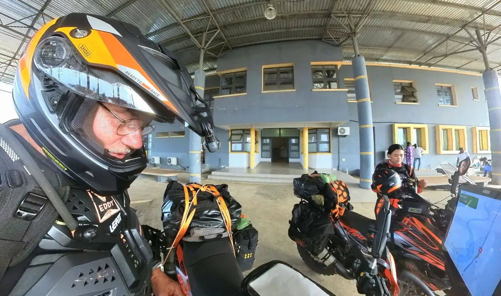 A wide angle view under the open sided canopy of a border post, Eddy in the foreground in helmet and chest armour with his visor up, both loaded motorcycles behind him on the concrete, and the grey blue immigration building with yellow trimmed windows beyond.