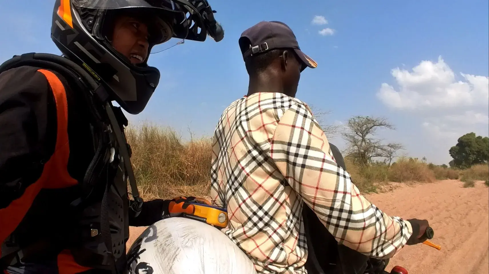 Seen from just behind, Max rides pillion in helmet and chest armour while a local man in a checked shirt and dark cap rides her loaded motorcycle down a red dirt track lined with dry grass and thorn trees.