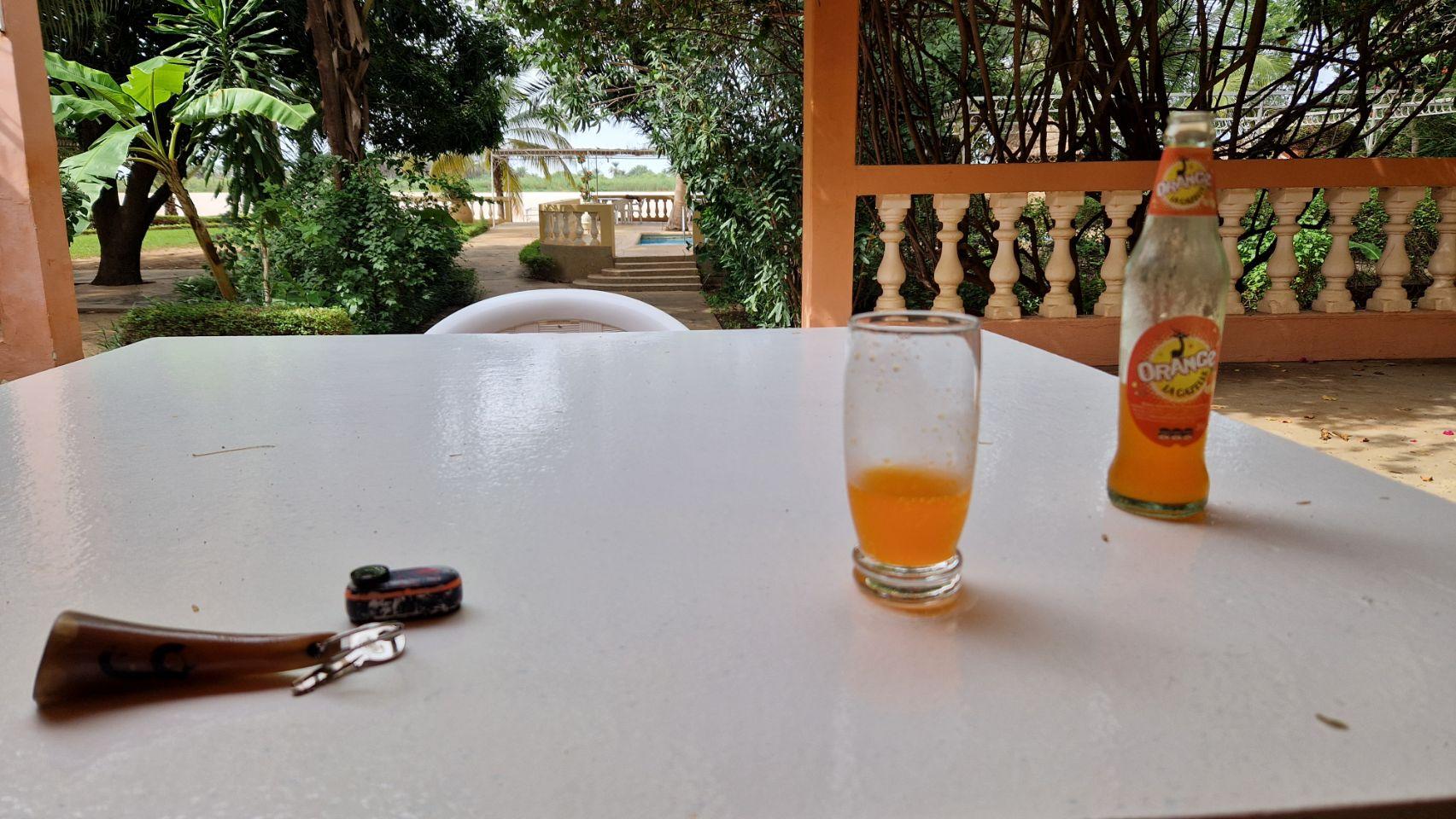 A glass of orange soda and its bottle sit on a table on a shaded porch, motorcycle keys nearby.