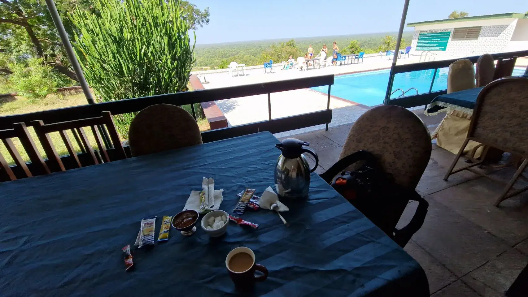 A table on a shaded terrace with a vacuum jug, a sugar bowl and instant coffee sachets, looking out past a swimming pool to bush stretching to the horizon.