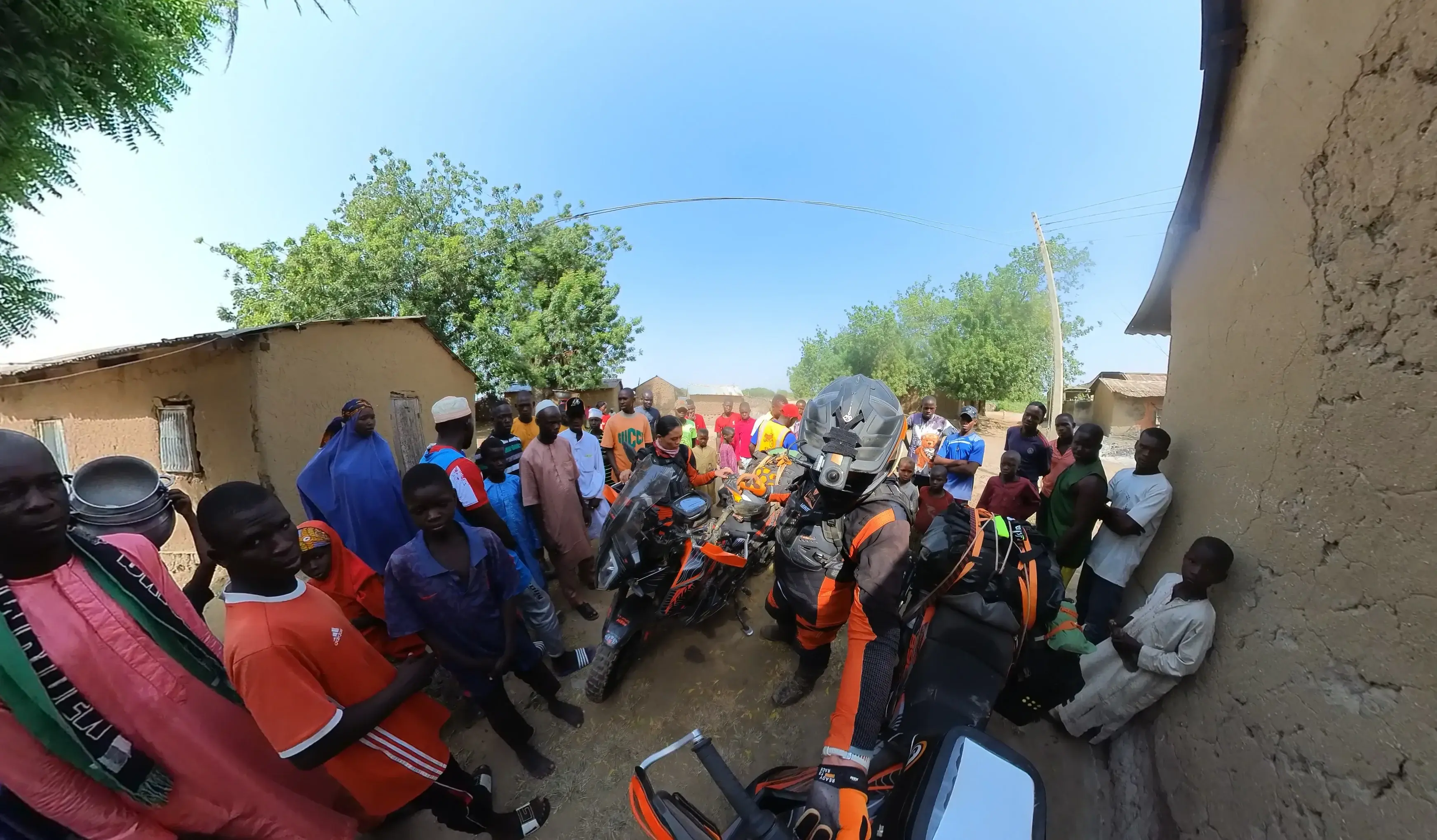 A large crowd of people gathered closely around two loaded motorcycles in a village street on a bright day.