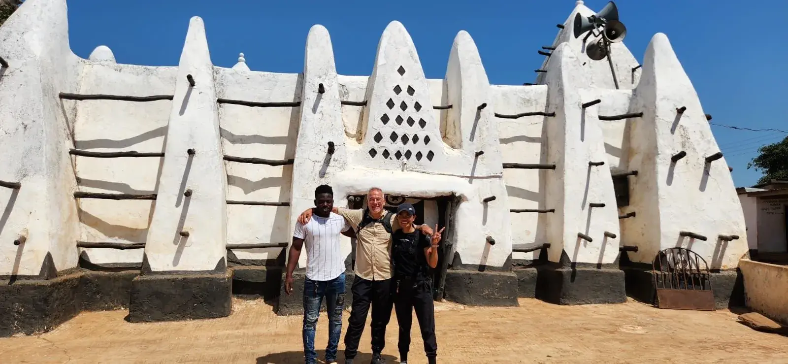 A whitewashed mosque built of mud and timber, its buttressed towers studded with protruding wooden beams, standing against a deep blue sky. Three people stand in the shade of the doorway.