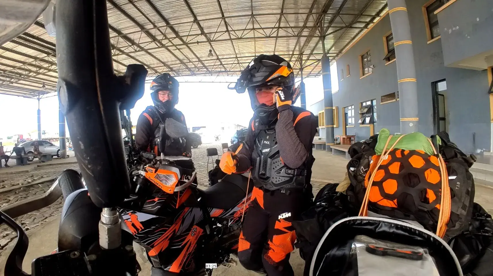 Two loaded orange and black adventure motorcycles parked under a corrugated steel canopy at a border post, luggage and dry bags stacked to one side, Max standing between them in riding armour giving a thumbs up.
