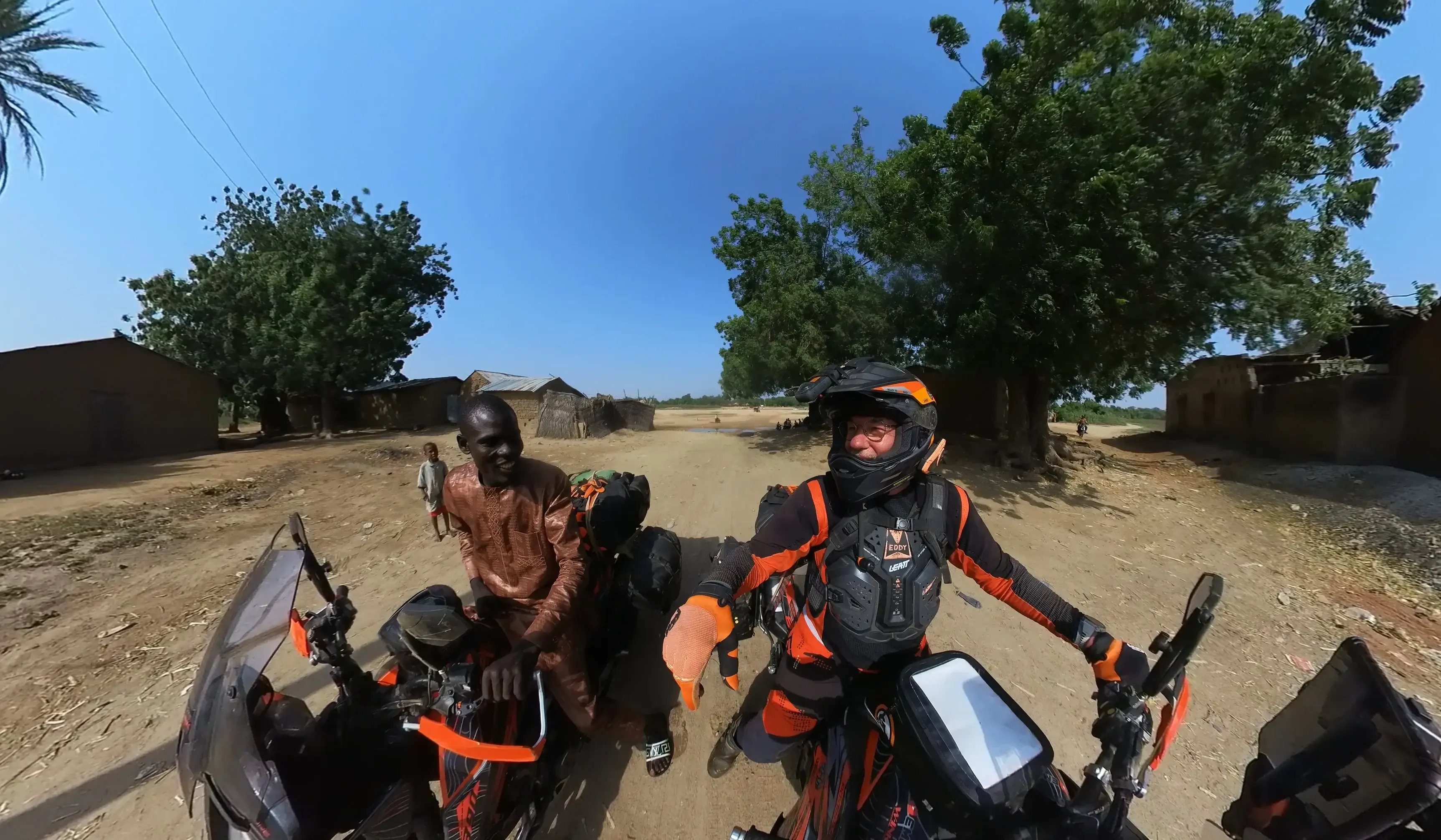 A rider on a loaded motorcycle stopped under a large tree, talking to a man standing beside the bike.