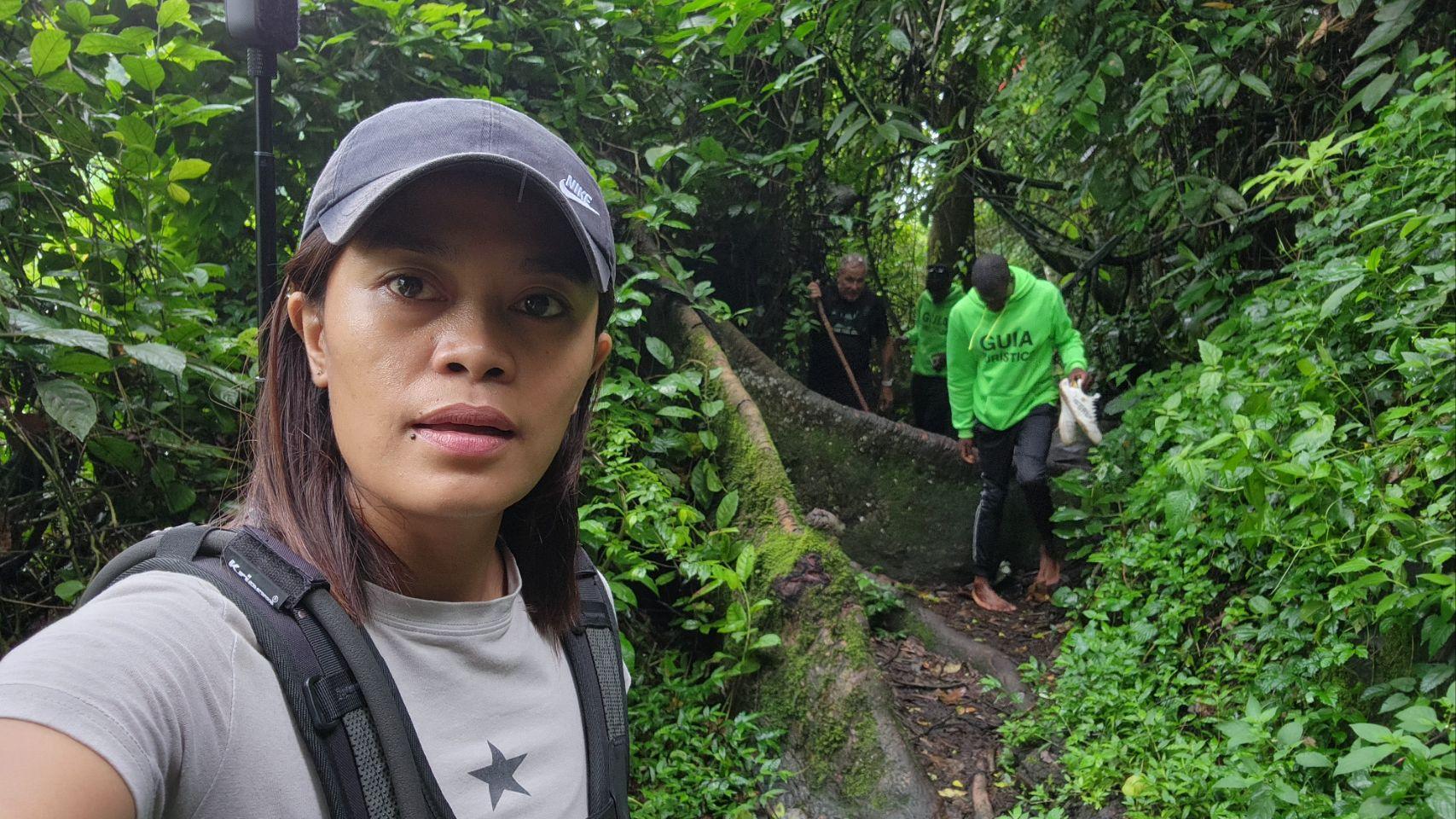 Max takes a selfie on a muddy jungle trail as two guides in bright green hoodies and Eddy, using a walking stick, follow behind.