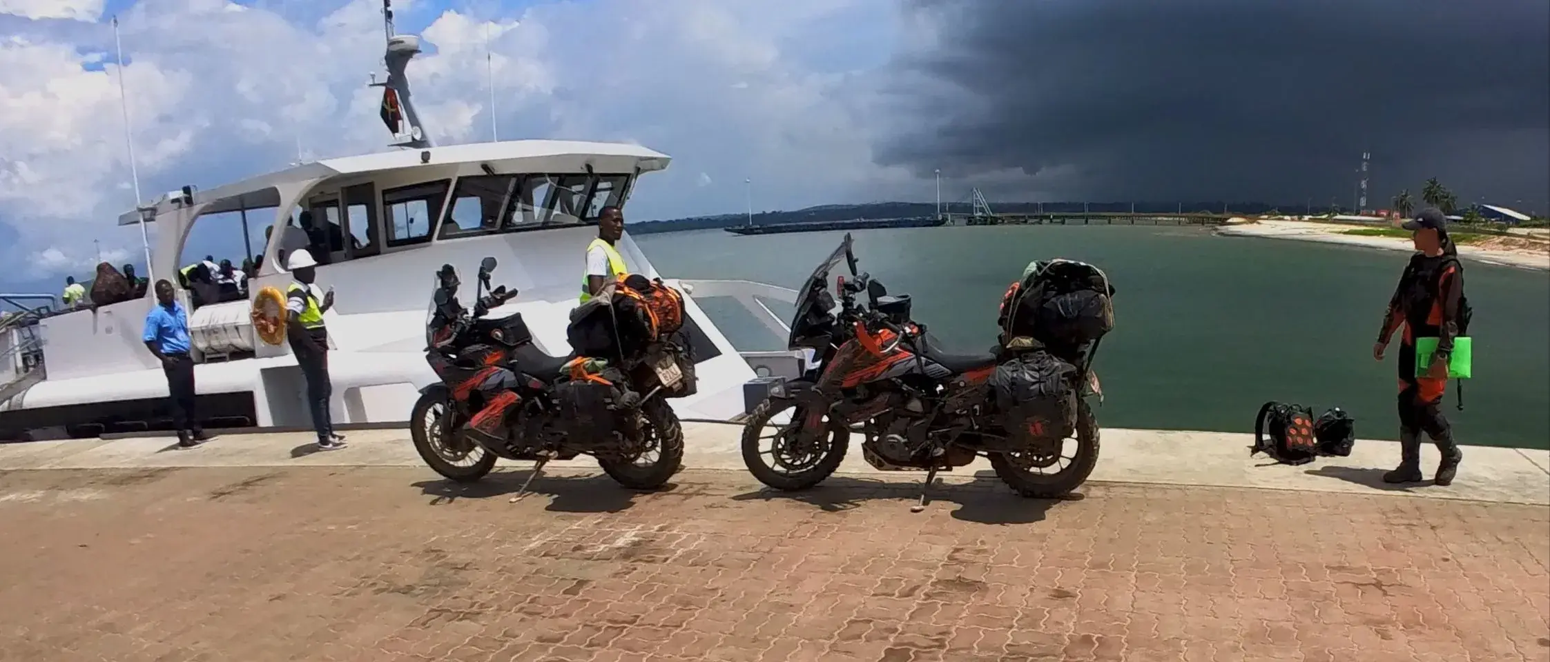 Two loaded KTM adventure motorcycles standing on a concrete quay in Cabinda, a white passenger catamaran moored behind them and a storm front sitting over the water.