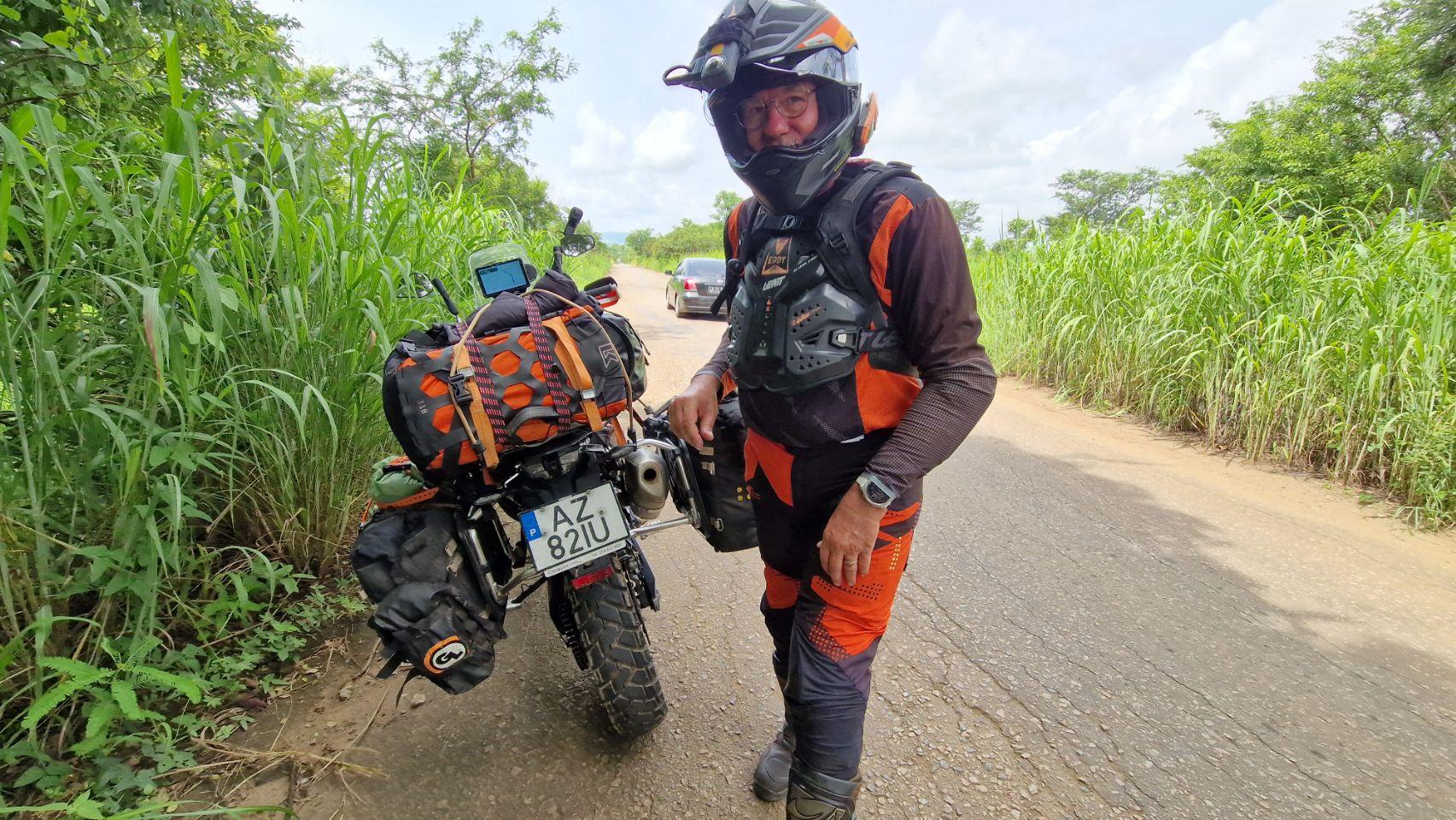 Eddy stands beside his fully loaded motorcycle on a smooth paved road cutting through green farmland in Angola.