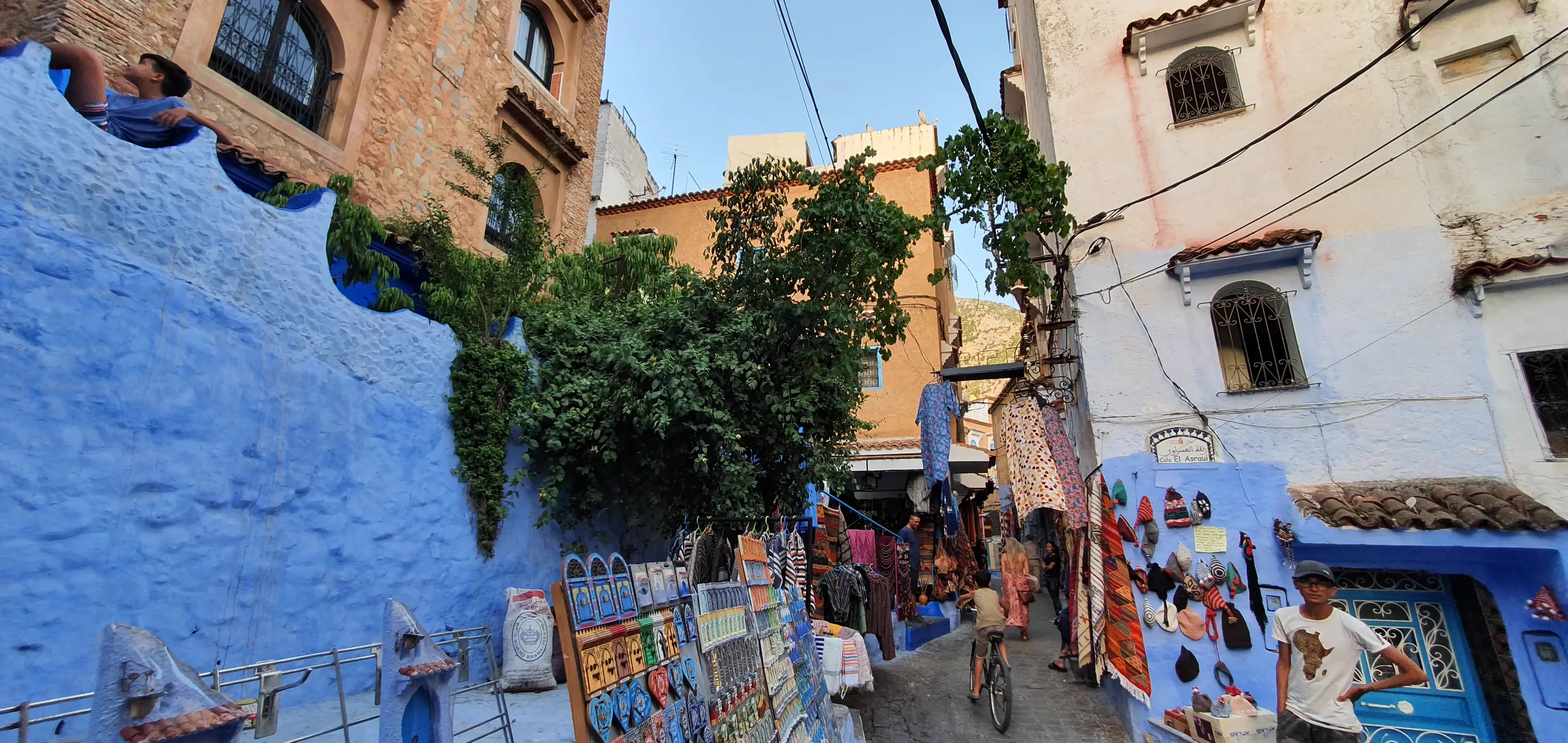 A narrow market street washed in blue, with rugs, paintings and painted goods hung out along both walls.