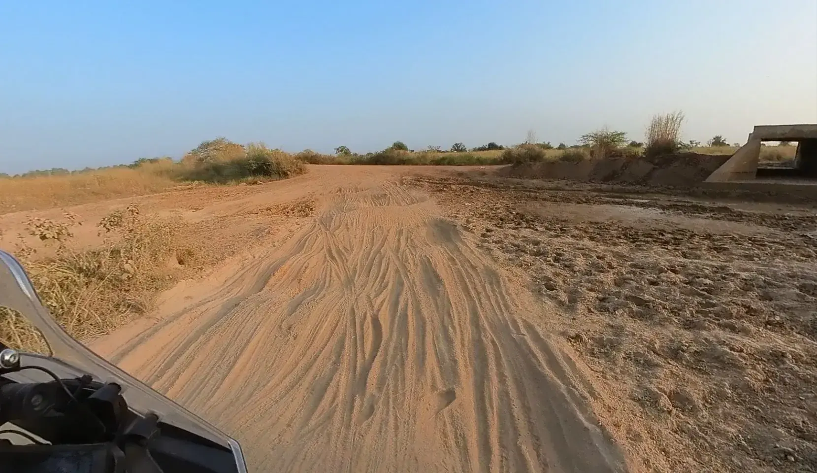 A rider's view over the handlebars along a deeply rutted track of soft orange sand, dry grass and scrub to one side, churned dark mud and a concrete culvert to the other, under a hazy blue sky.