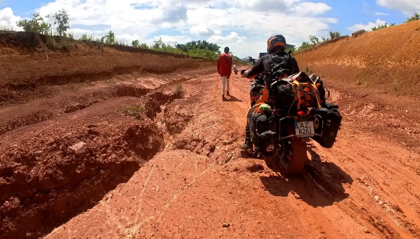 A loaded adventure motorcycle standing in a deeply rutted, churned red clay track, a person walking ahead down the same road.