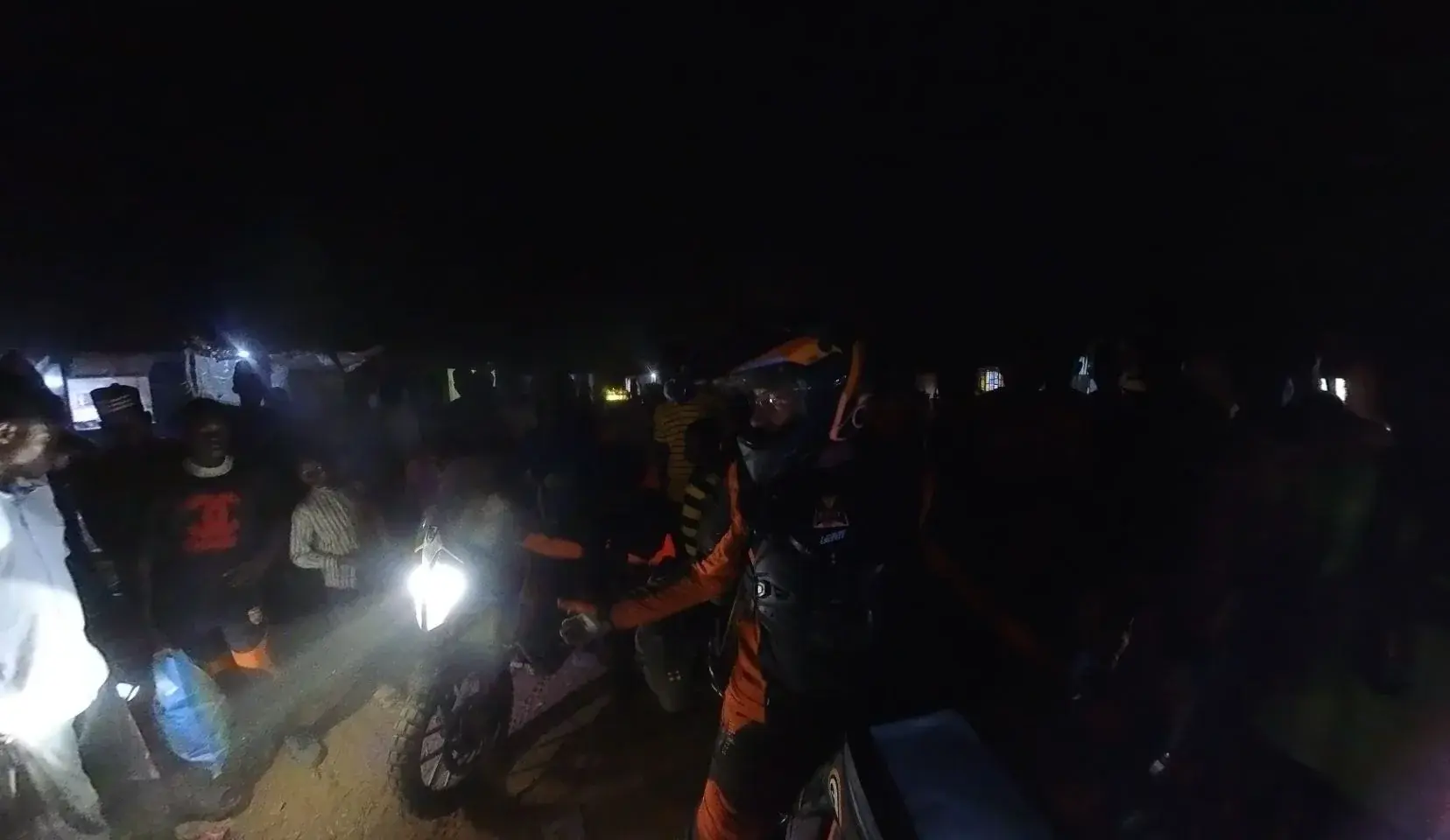 A night scene lit only by a motorcycle headlight. Eddy sits on his bike still helmeted with the visor down, surrounded by a crowd of people reduced mostly to silhouettes, with faint warm lights from buildings far behind.