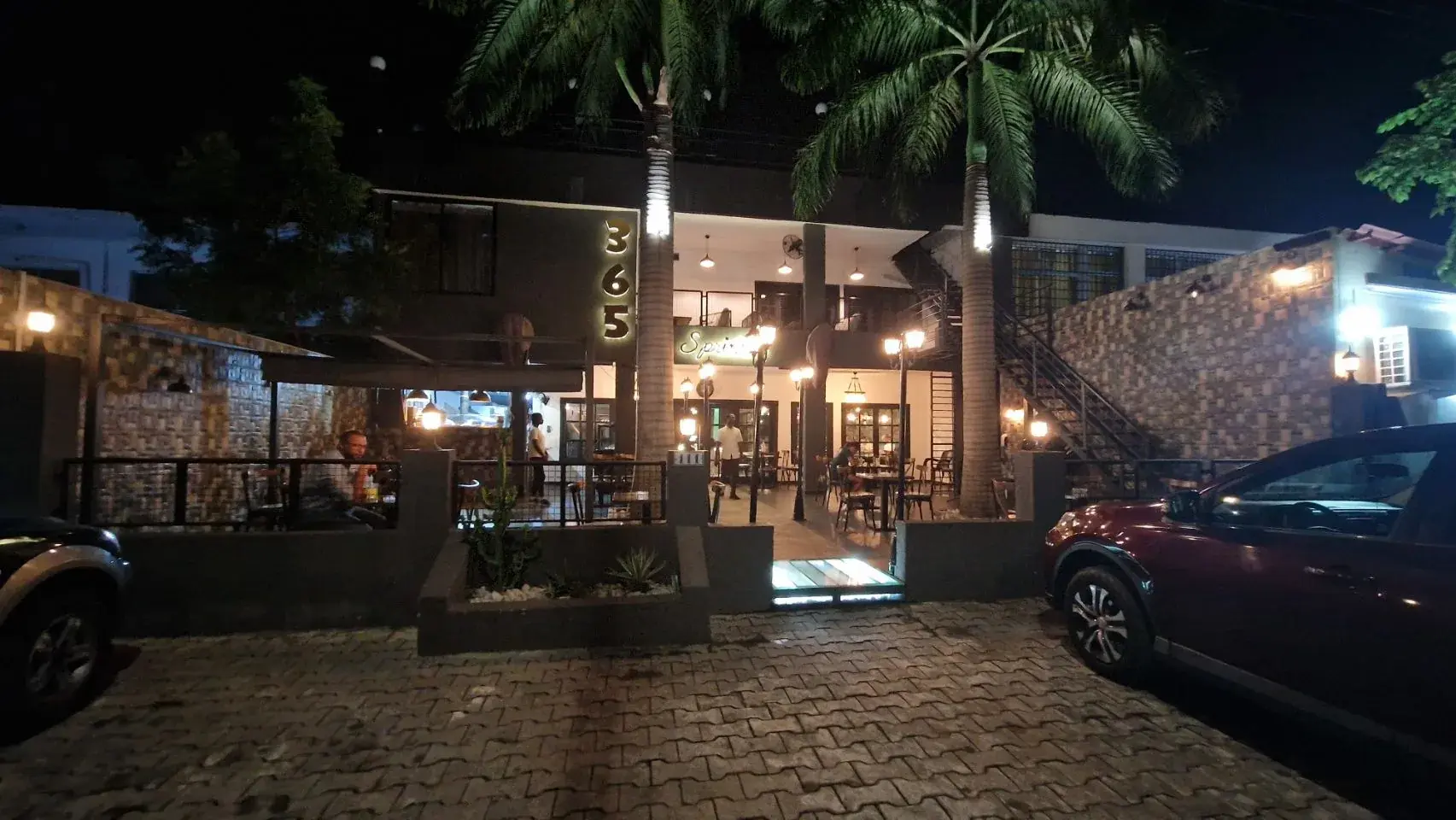 A restaurant lit up at night, its illuminated sign reading 365 above an open fronted ground floor with warm pendant lights, two floodlit palm trees framing the entrance and cars parked on the brick driveway.