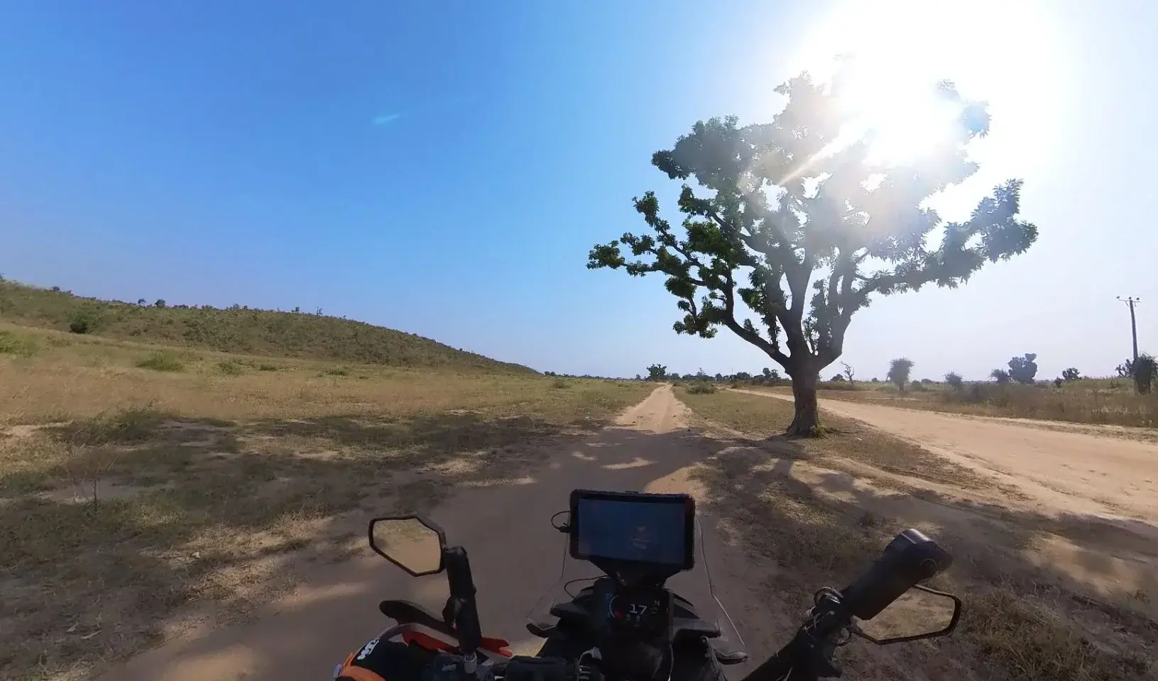 Handlebar view of a sandy two track dirt road running toward a single leafy tree backlit by the sun, dry grassland either side and a low hill on the left under a deep blue sky.