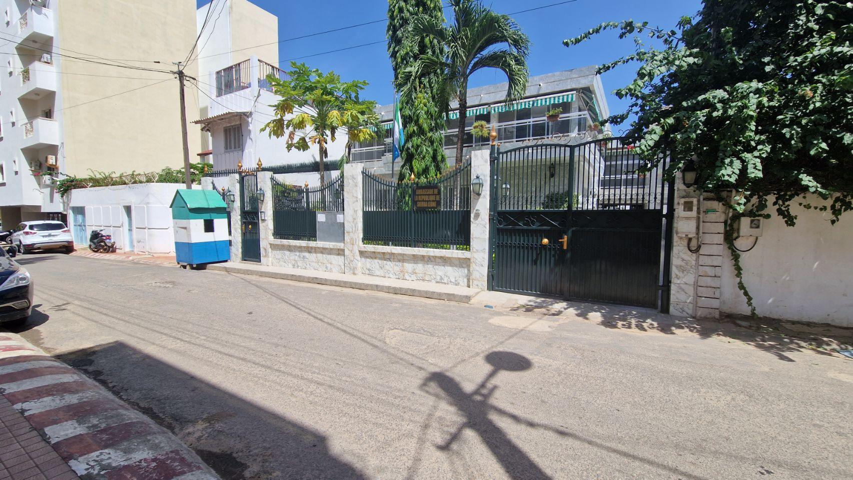 A green metal gate and marble-topped wall mark the entrance to an embassy building on a quiet Dakar street.