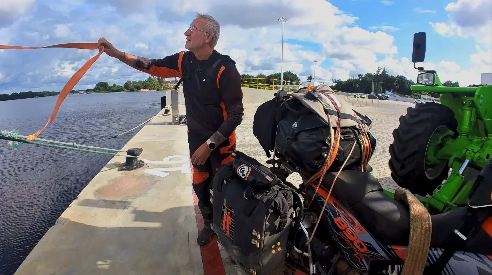 Eddy stands on the quay in Soyo holding out an orange lifting strap, his loaded bike beside him and the arm of a green telehandler at the edge of the frame.