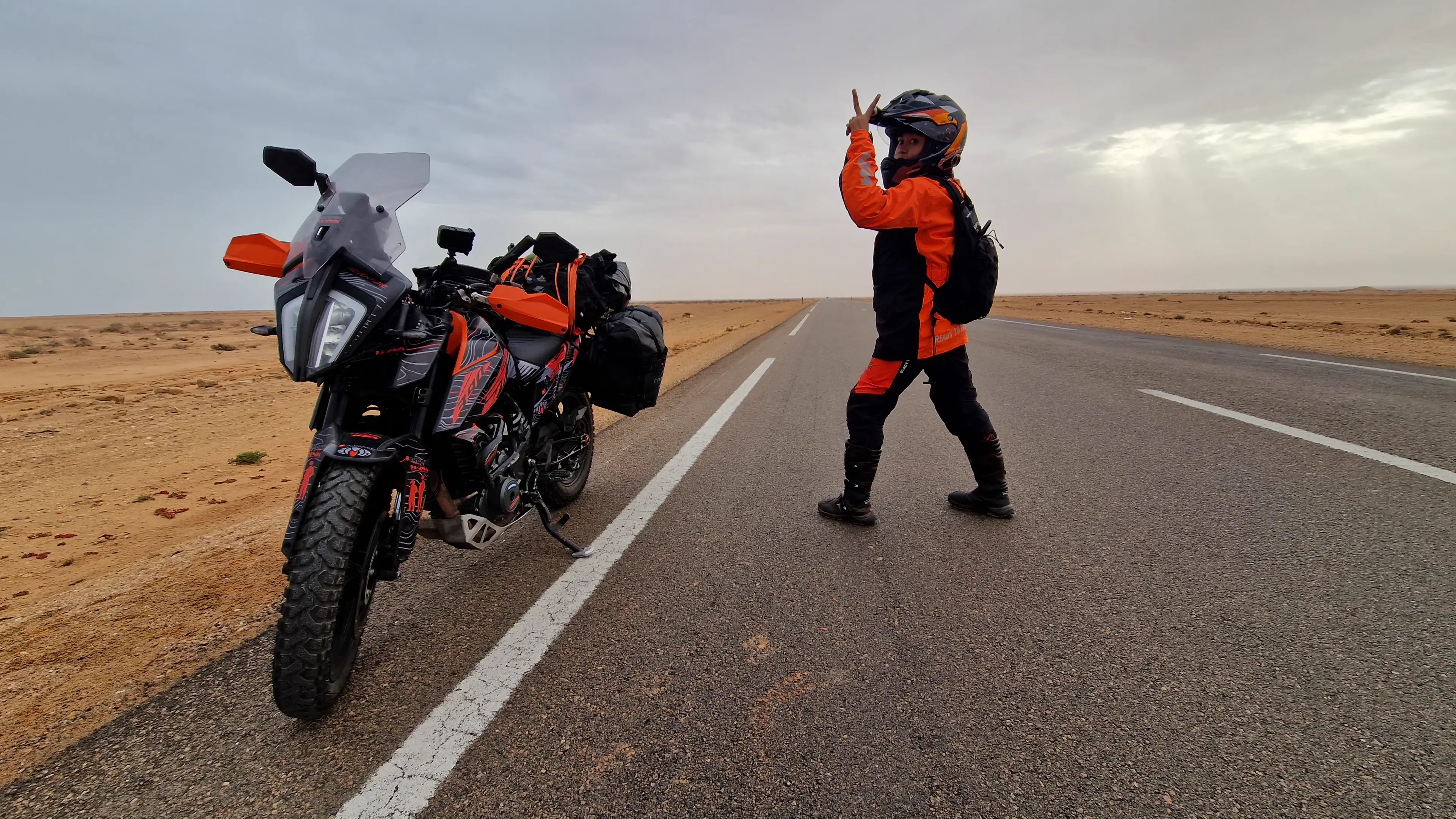 A rider in orange gear striding across an empty desert highway with both arms raised, a heavily loaded motorcycle parked on the shoulder behind, flat scrub running to the horizon under a grey sky.