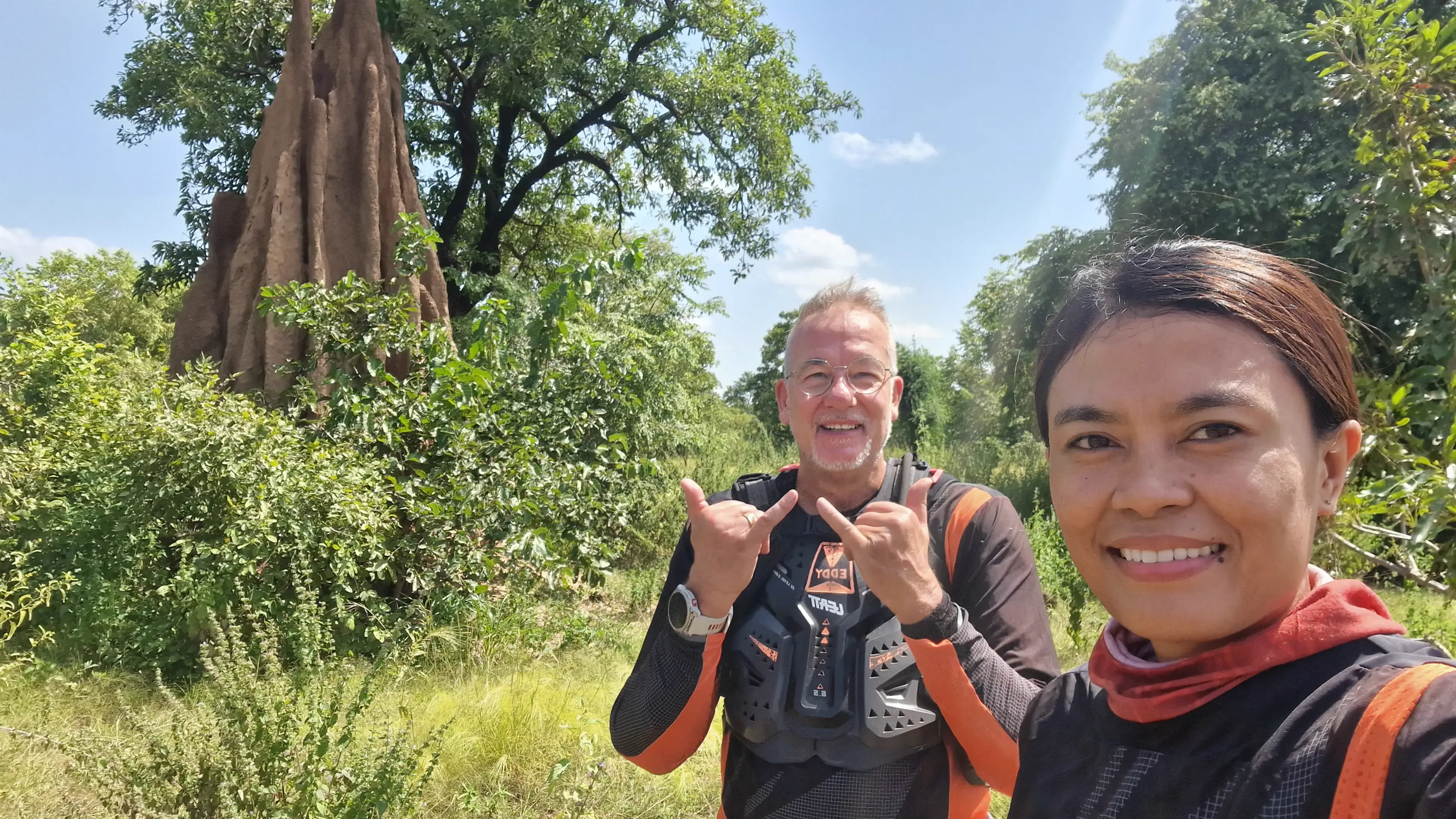 Max taking a selfie of the two of them in riding gear on a green bush track, Eddy grinning behind in body armour with a tall termite mound and trees beyond.