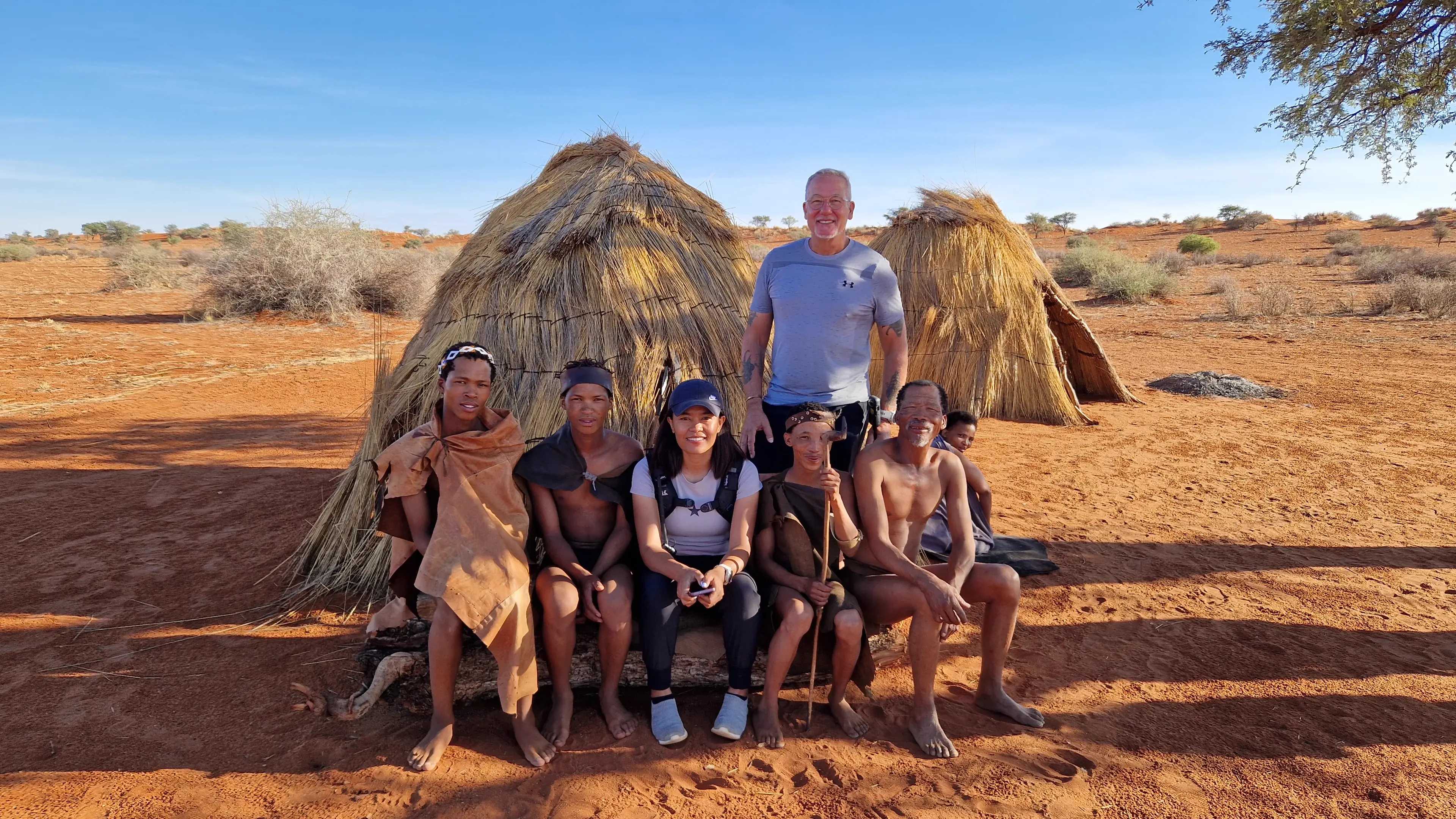 Eddy standing and Max seated among a family group in front of two thatched grass shelters on red Kalahari sand, everyone facing the camera in bright midday sun.