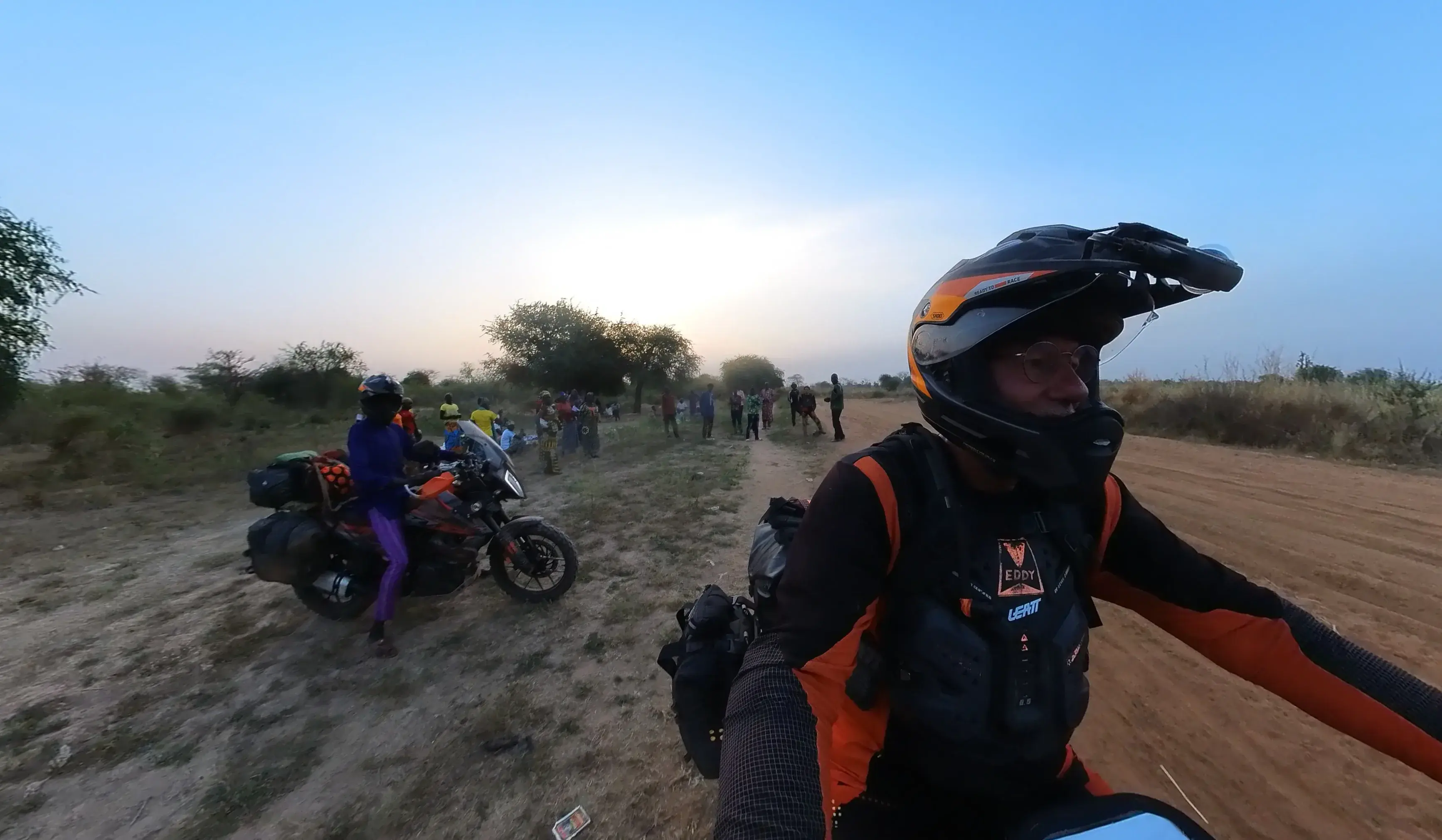 Eddy on his motorcycle at dusk on a dirt track, name-tagged body armour visible, with Max and a gathered crowd of onlookers further down the track in the low sun.